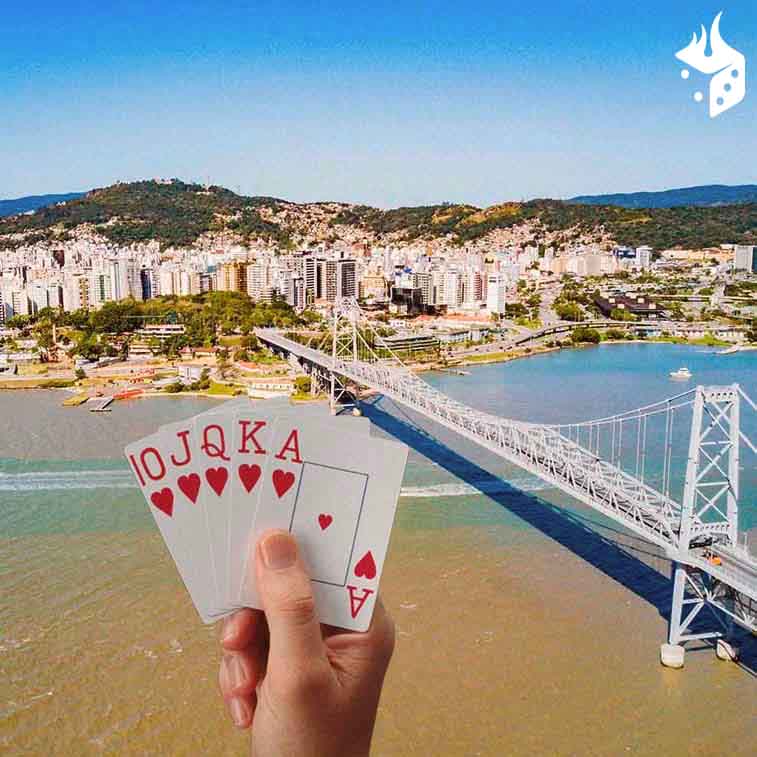floripa no poker Ponte Hercílio Luz em evidência na imagem com uma mão segurando cartas de poker